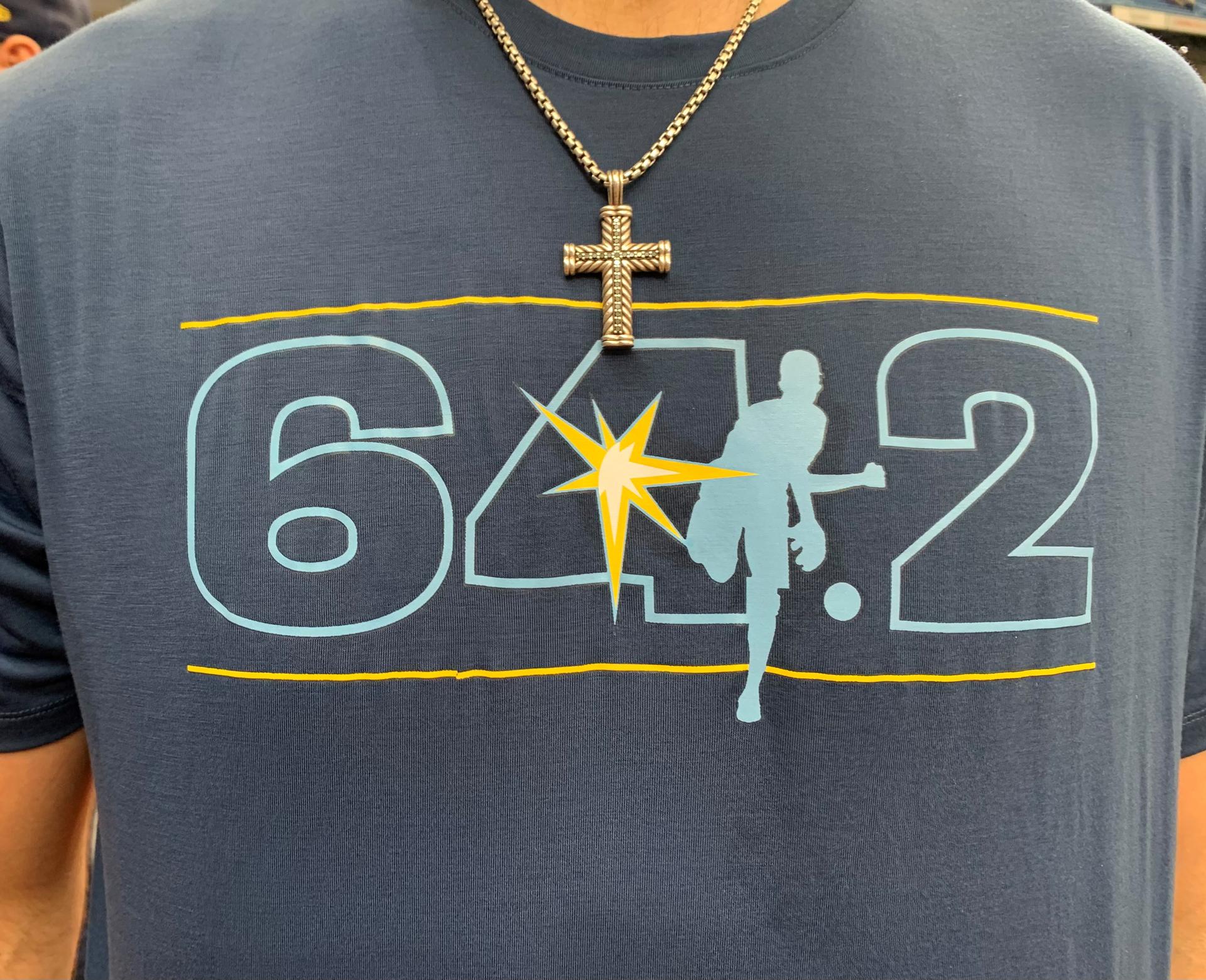 Close-up photo of a blue T-shirt that reads: 64.2. The Rays' sunburst logo is inside the ''4,'' with a pitcher throwing a ball representing the decimal.