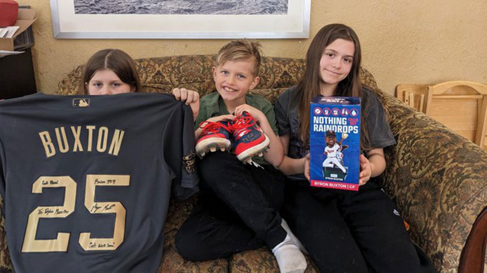 Three children sit on a sofa. One holds a signed jersey, one a pair of cleats and one a box
