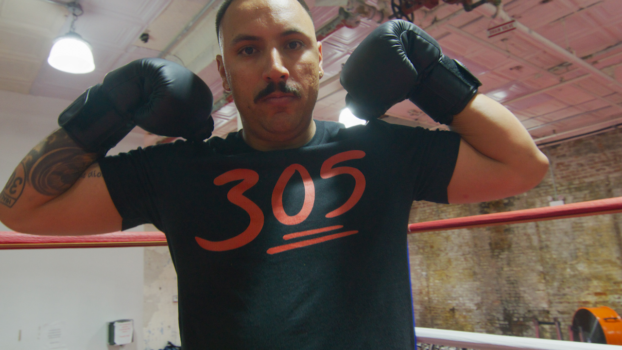 A man wearing boxing gloves flexes his arms while wearing a black T-shirt with the number 305 on it